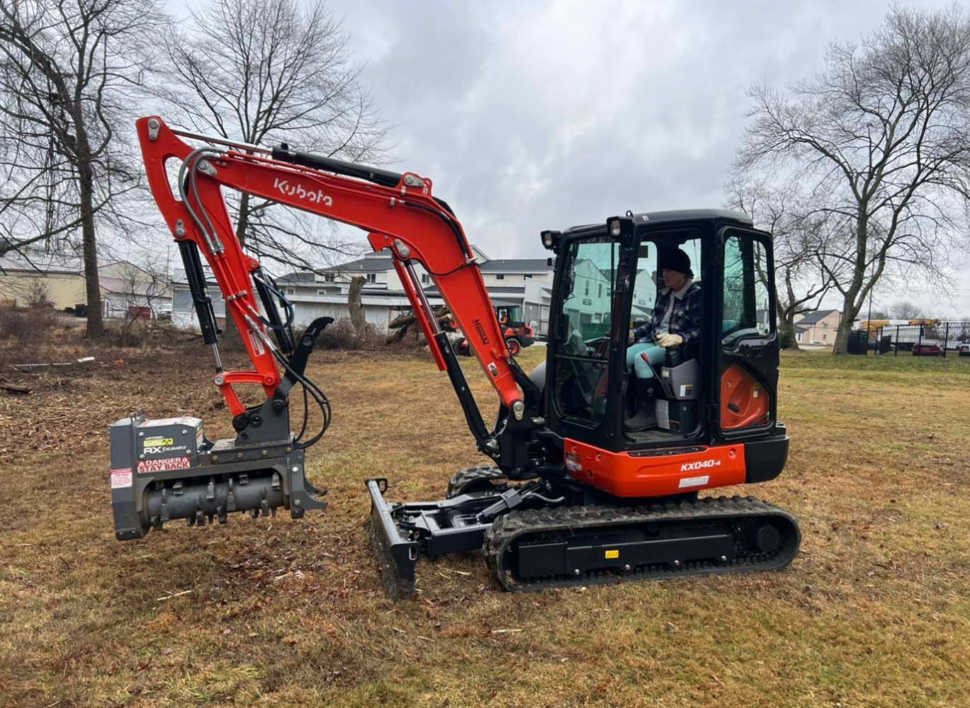 Orange Kubota excavator with a forestry mulcher attachment, parked on a grassy field under a cloudy sky.
