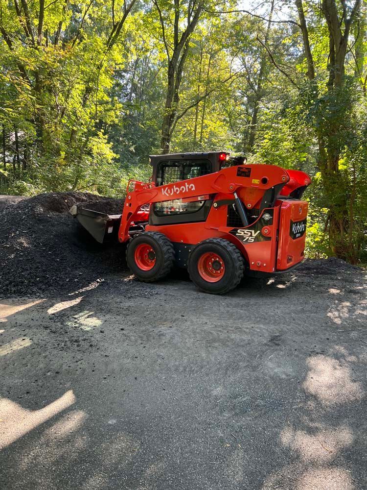 A bright orange Kubota skid-steer loader pushing a pile of dark gravel in a wooded area on a sunny day.