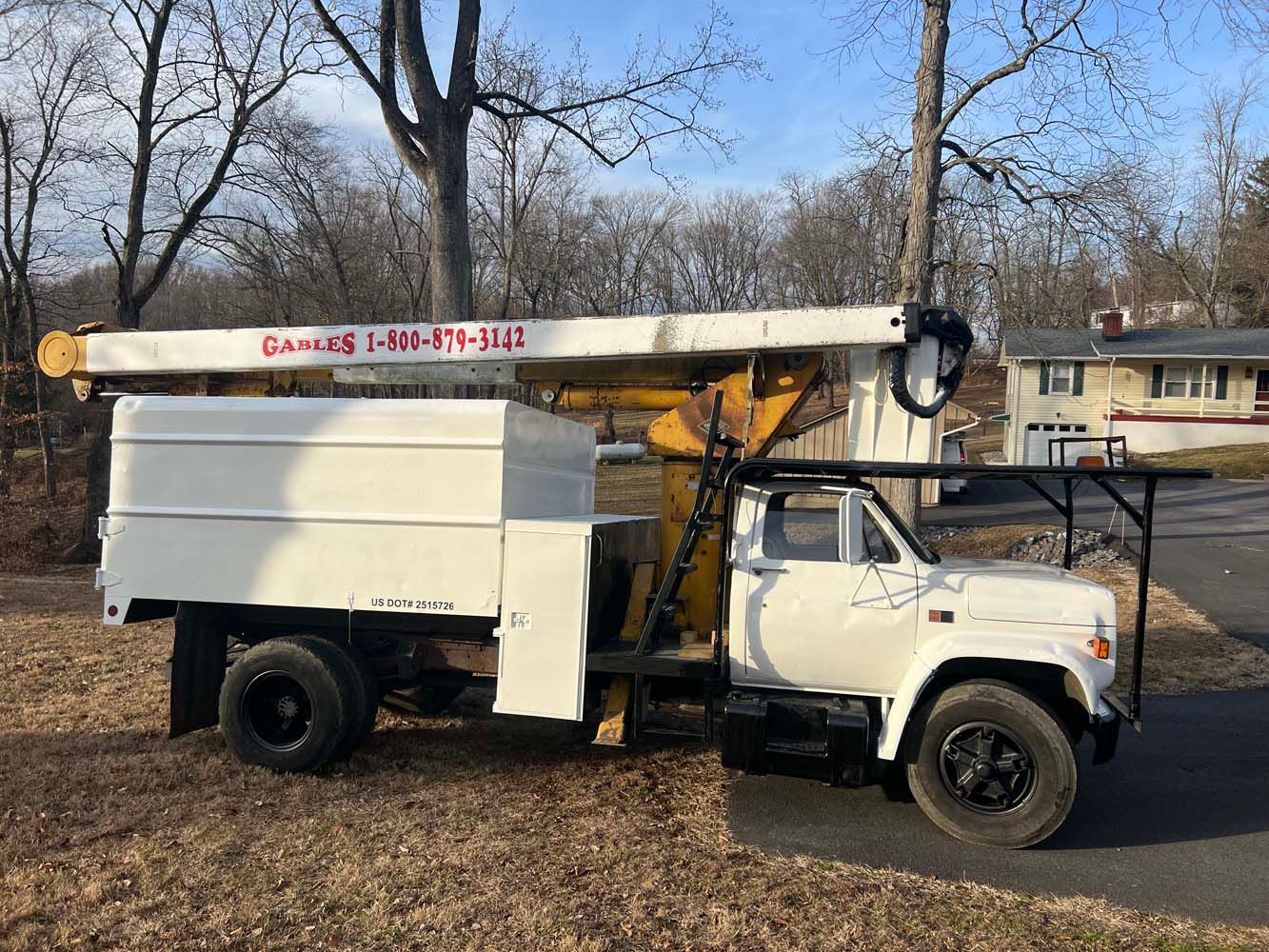 A white utility bucket truck parked on a grassy patch next to a residential driveway in a wooded neighborhood.