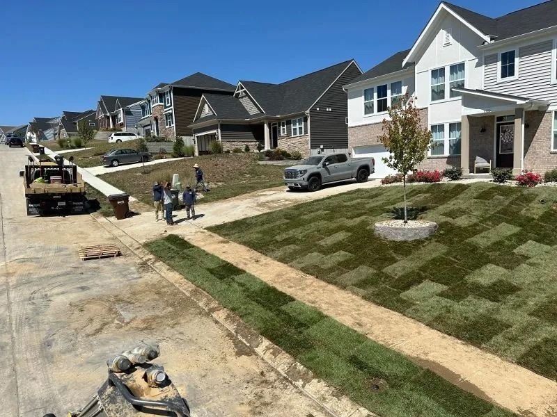 Suburban street with new homes, parked trucks, workers on a driveway, and a freshly sodded front yard.