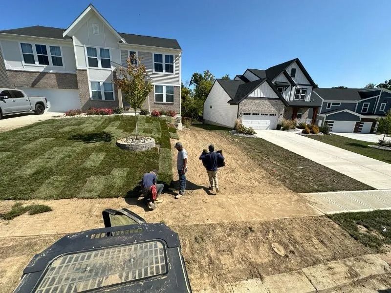 Workers on a suburban lawn near a driveway, with houses and a truck in the background.