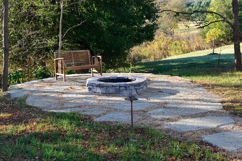Stone fire pit beside a wooden bench on a grassy park path, surrounded by trees and sunlight
