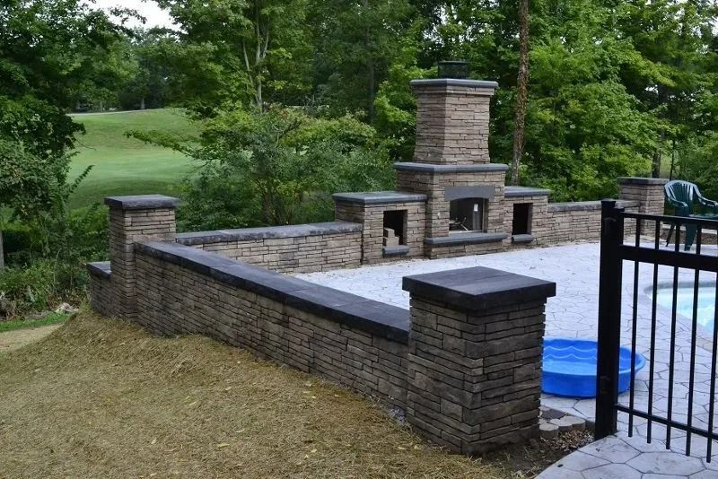 Outdoor stone patio with built-in fireplace, low wall, and black gate beside a blue covered pool