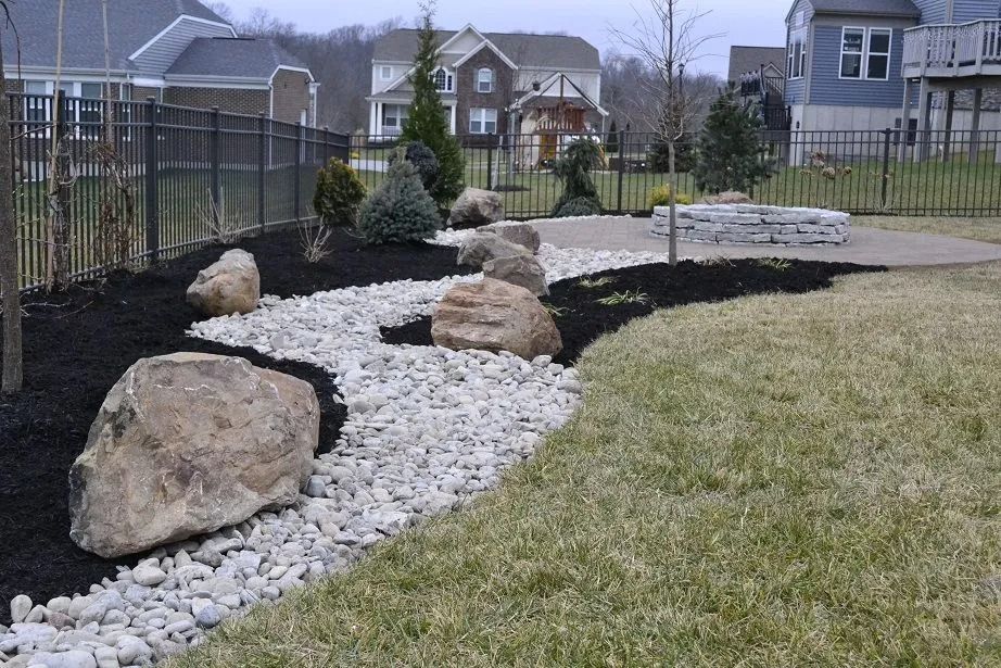 Rock garden with white stones and large boulders beside a lawn and fence in a suburban yard