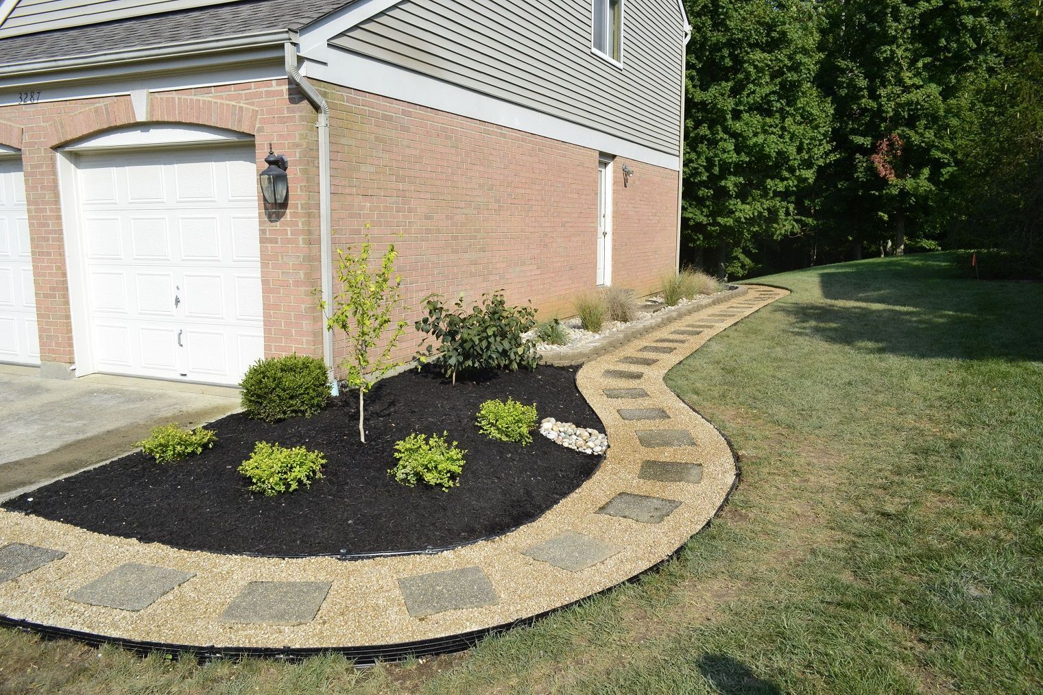 Curved stone walkway beside a brick house, lined with mulch and small shrubs, leading to a grassy yard.