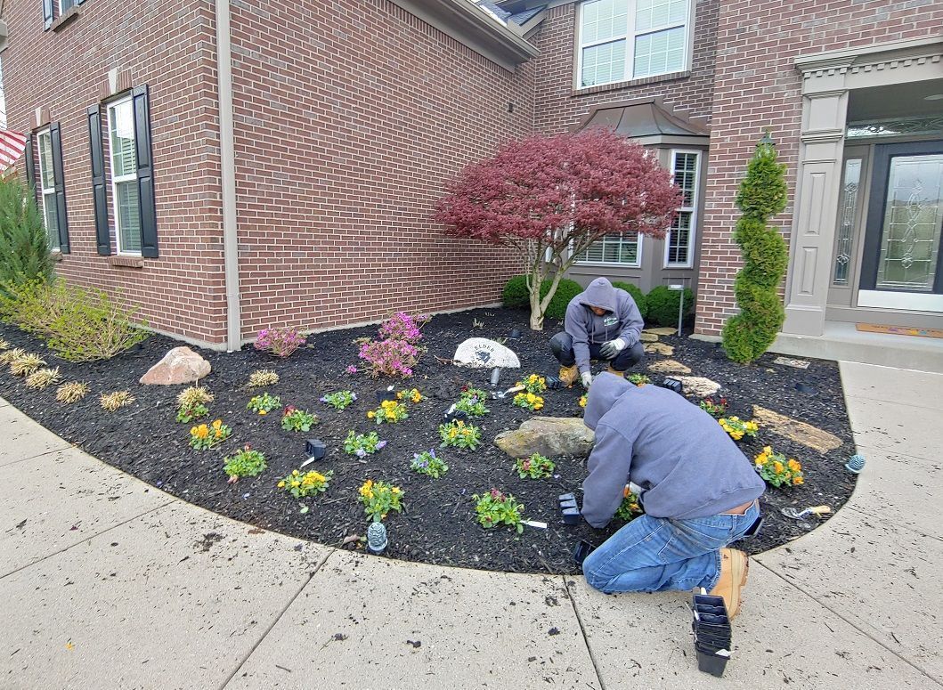 Two workers planting flowers in a curved garden bed beside a brick house