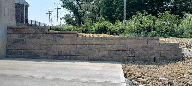 Stone retaining wall beside a paved driveway, with dirt and greenery in the background