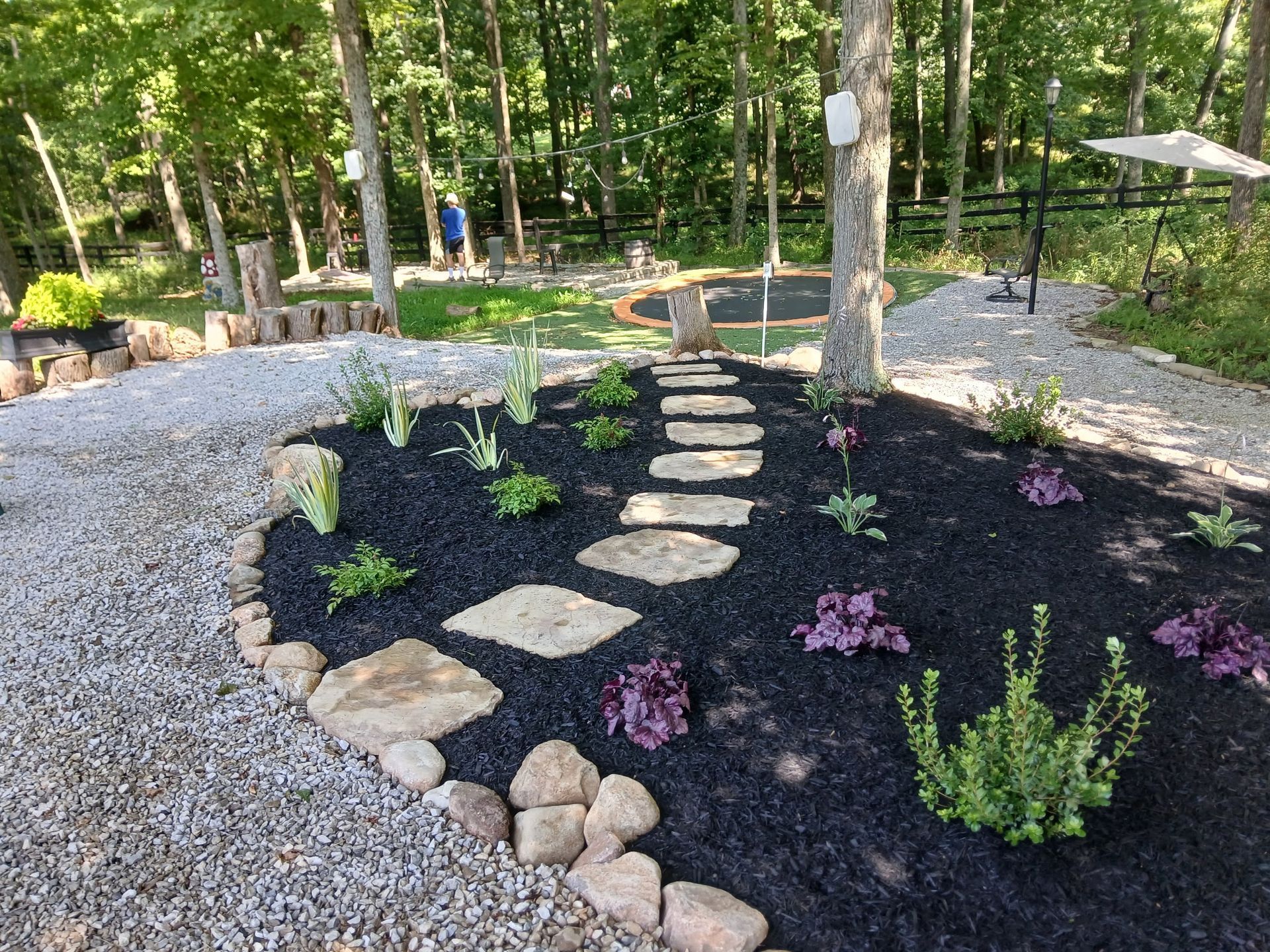 Garden path of stepping stones through a dark mulch bed with small plants, surrounded by gravel and trees