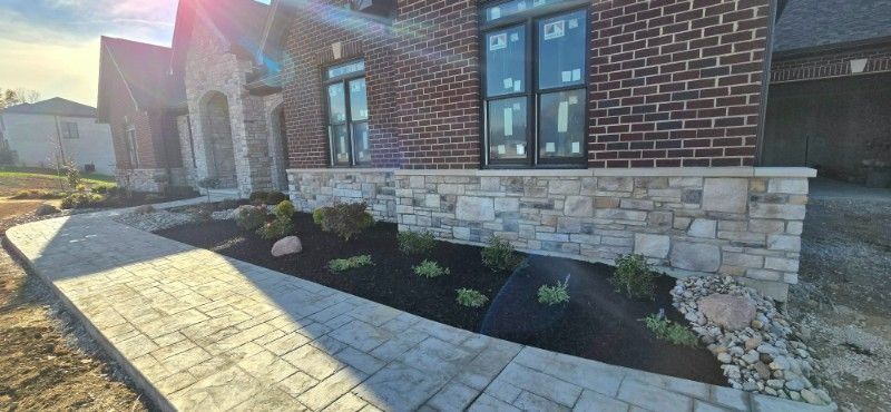 Brick house exterior with stone foundation, black mulch beds, and a sunlit paved walkway