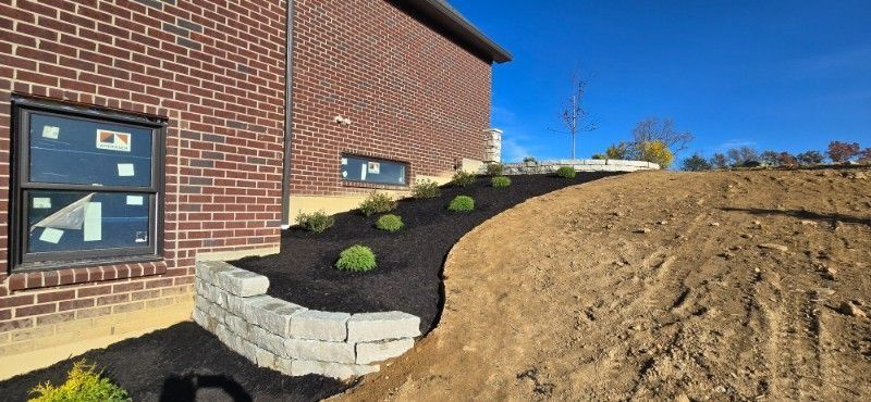 Brick house beside newly landscaped sloped yard with retaining wall, dark mulch, and young shrubs
