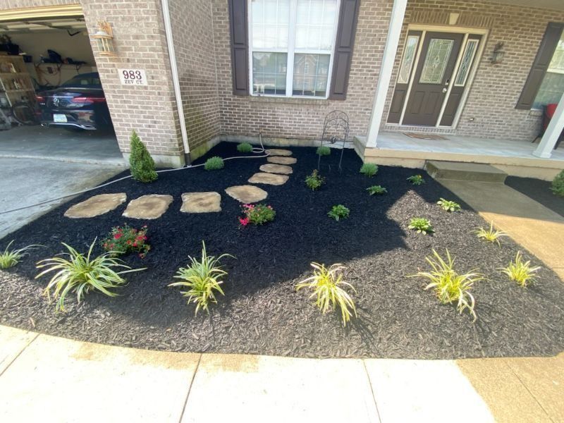 Front porch landscaping with black mulch, stepping stones, and small green plants by a brick house.