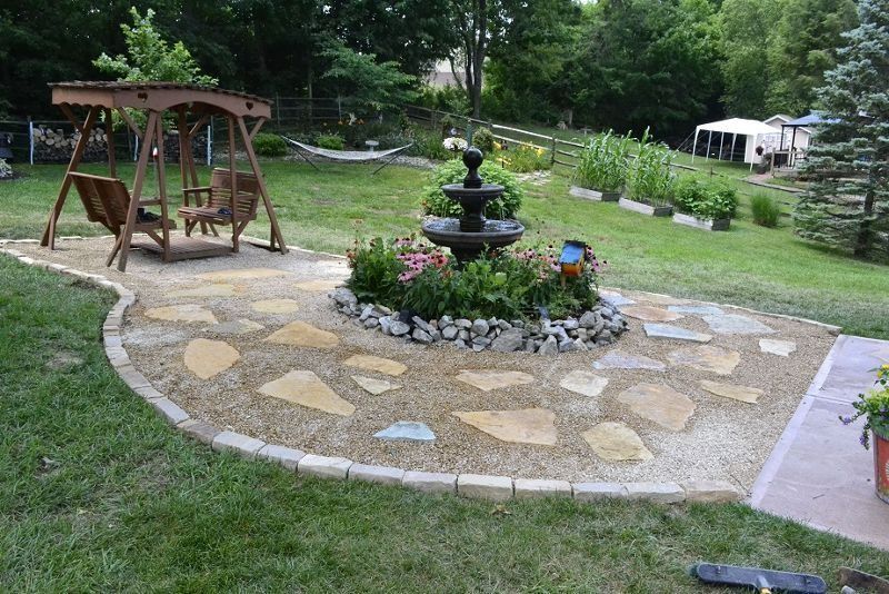 Garden patio with a stone path, flower planter fountain, and wooden swing bench surrounded by greenery