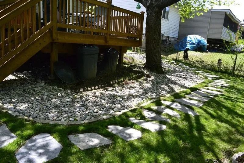 Stone path across a grassy yard beside a wooden deck and tree, with a blue-covered item in the background