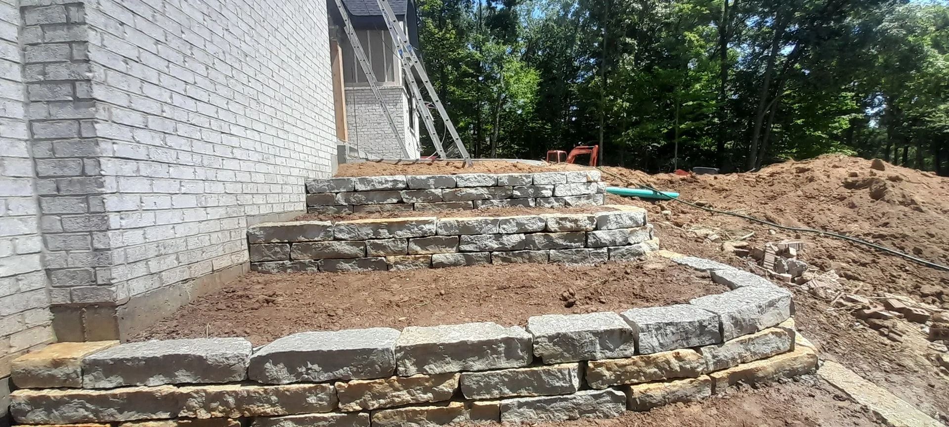 Stone retaining wall and steps under construction beside a white brick house.