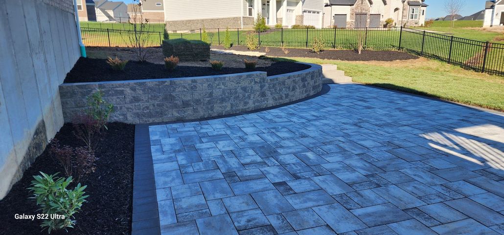 Stone patio and landscaped backyard with retaining wall, mulch beds, and neighboring houses in the background