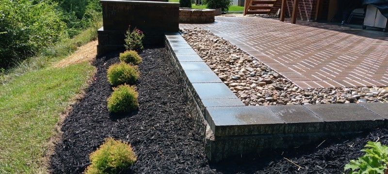 Modern landscaped garden bed with black mulch, small shrubs, and a raised stone border beside a walkway