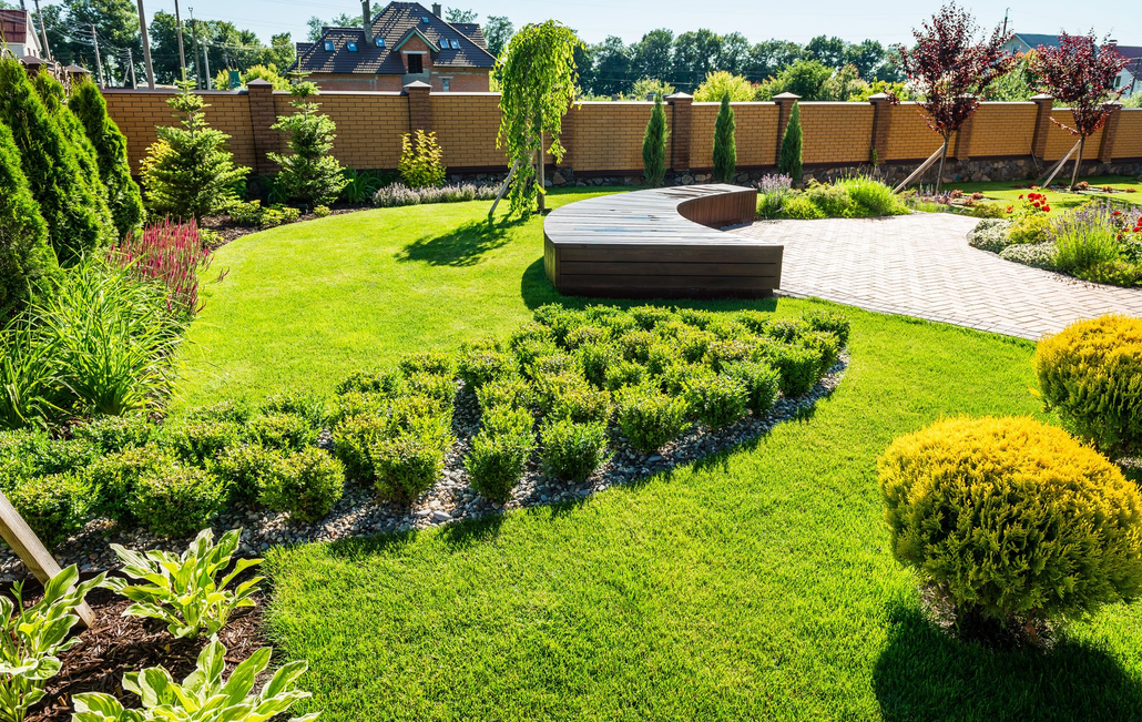 Sunny landscaped garden with curved lawn, stone seating, and flower beds behind a wooden fence