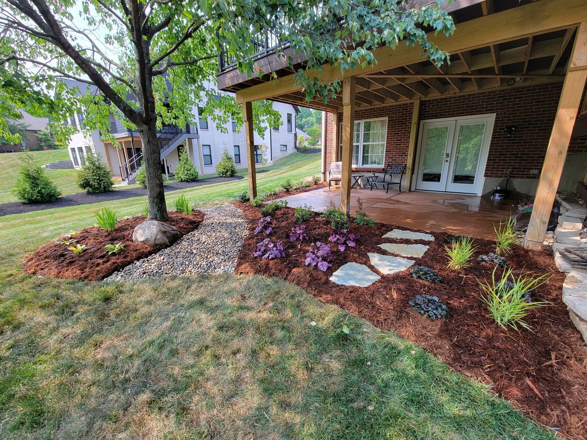 Wooded front yard with a covered porch, stepping stones, mulch beds, and a leafy tree beside the lawn