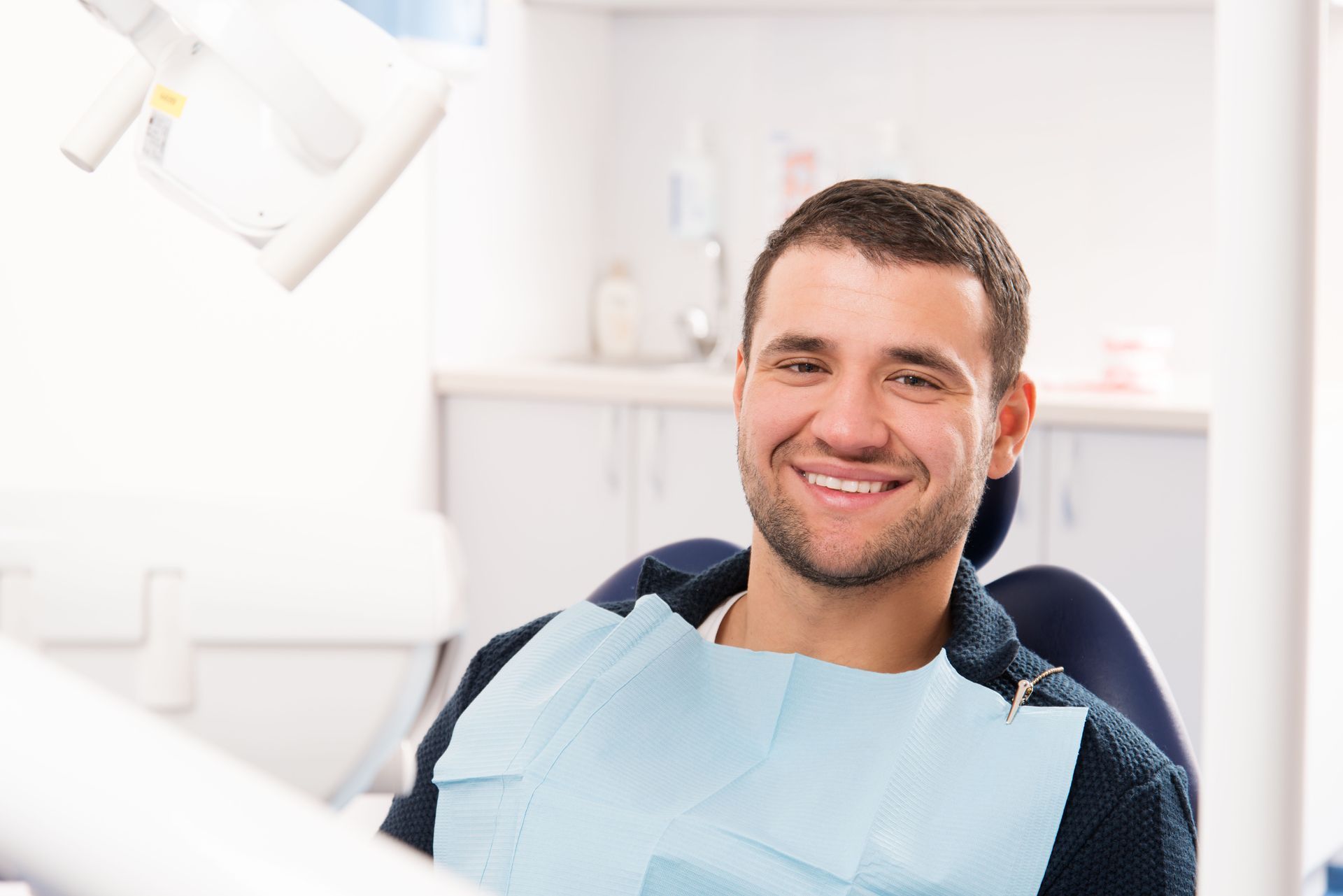 A smiling person sits in a dentist's chair wearing a light blue dental bib in a bright office.