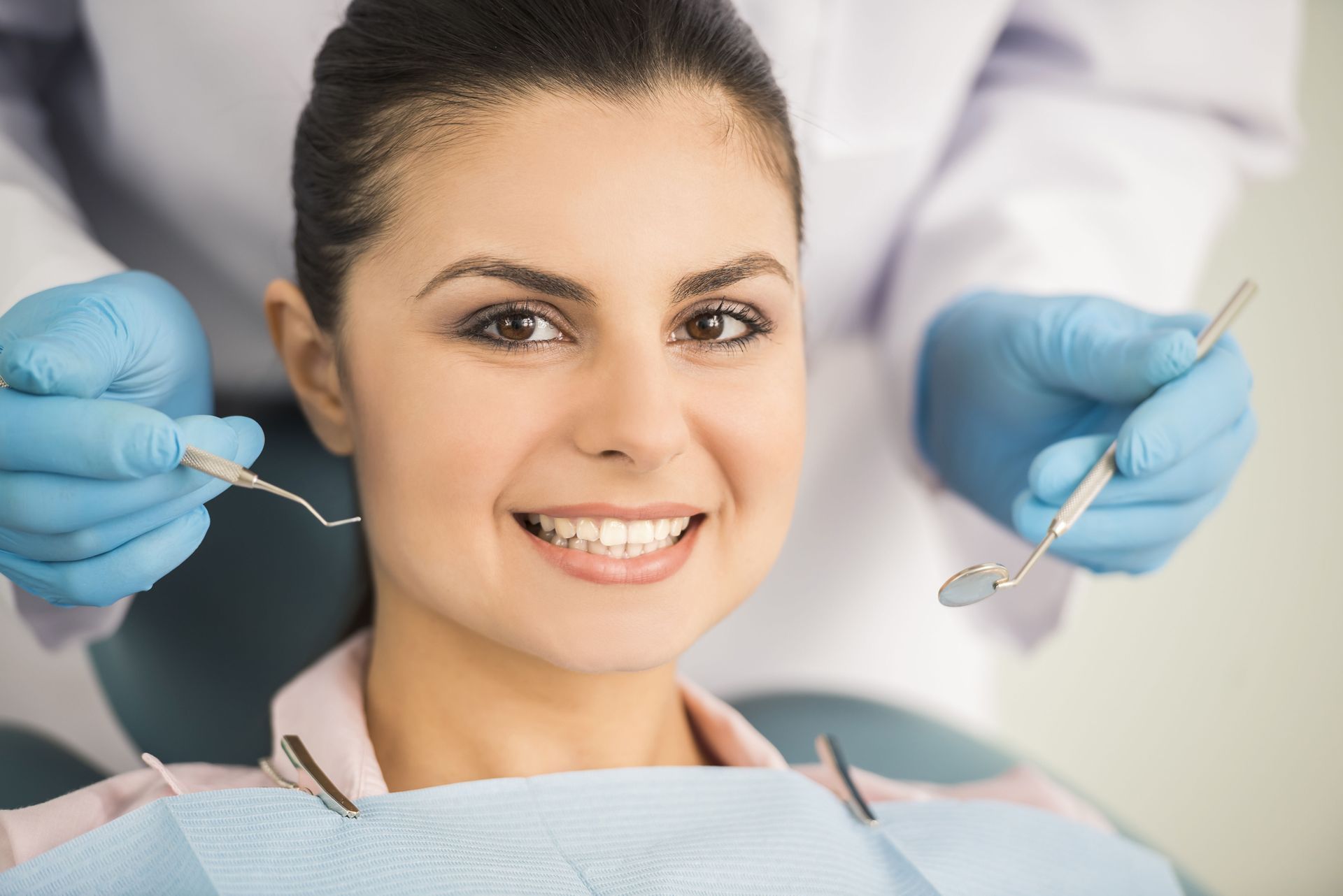 A smiling person sitting in a dental chair with a provider wearing blue medical gloves holding a dental mirror and probe.