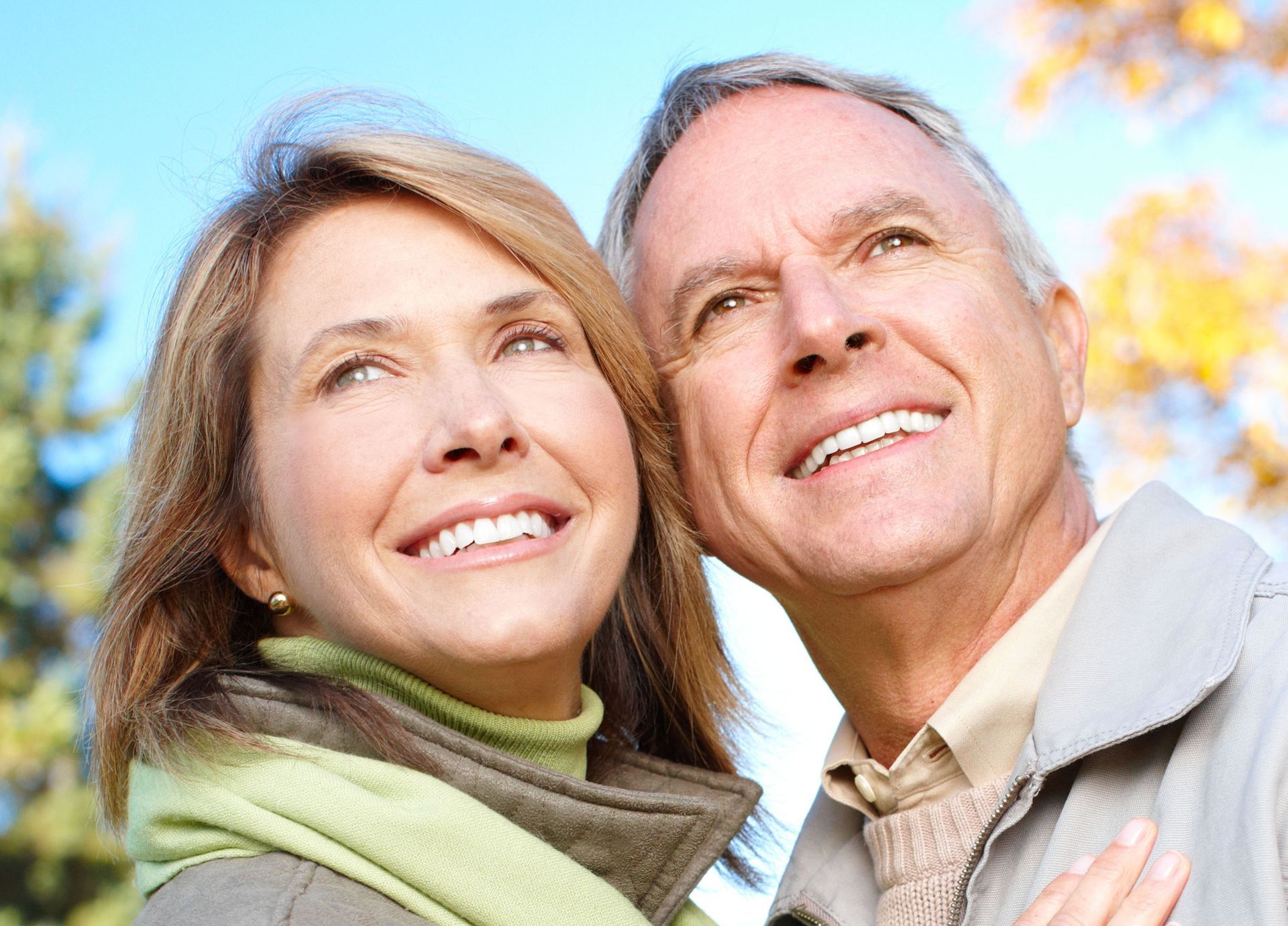 A smiling couple looks upward together outdoors against a background of trees and blue sky.