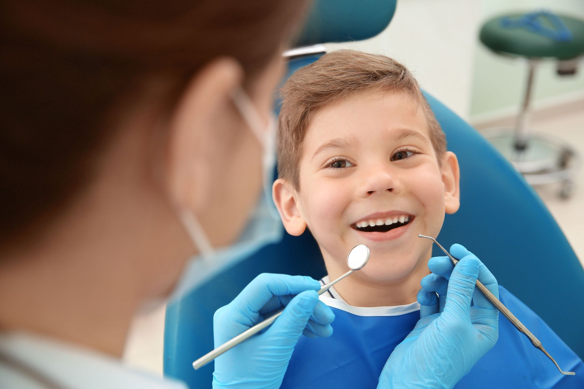 A dentist in blue gloves uses instruments to examine the mouth of a smiling patient sitting in a dental chair.