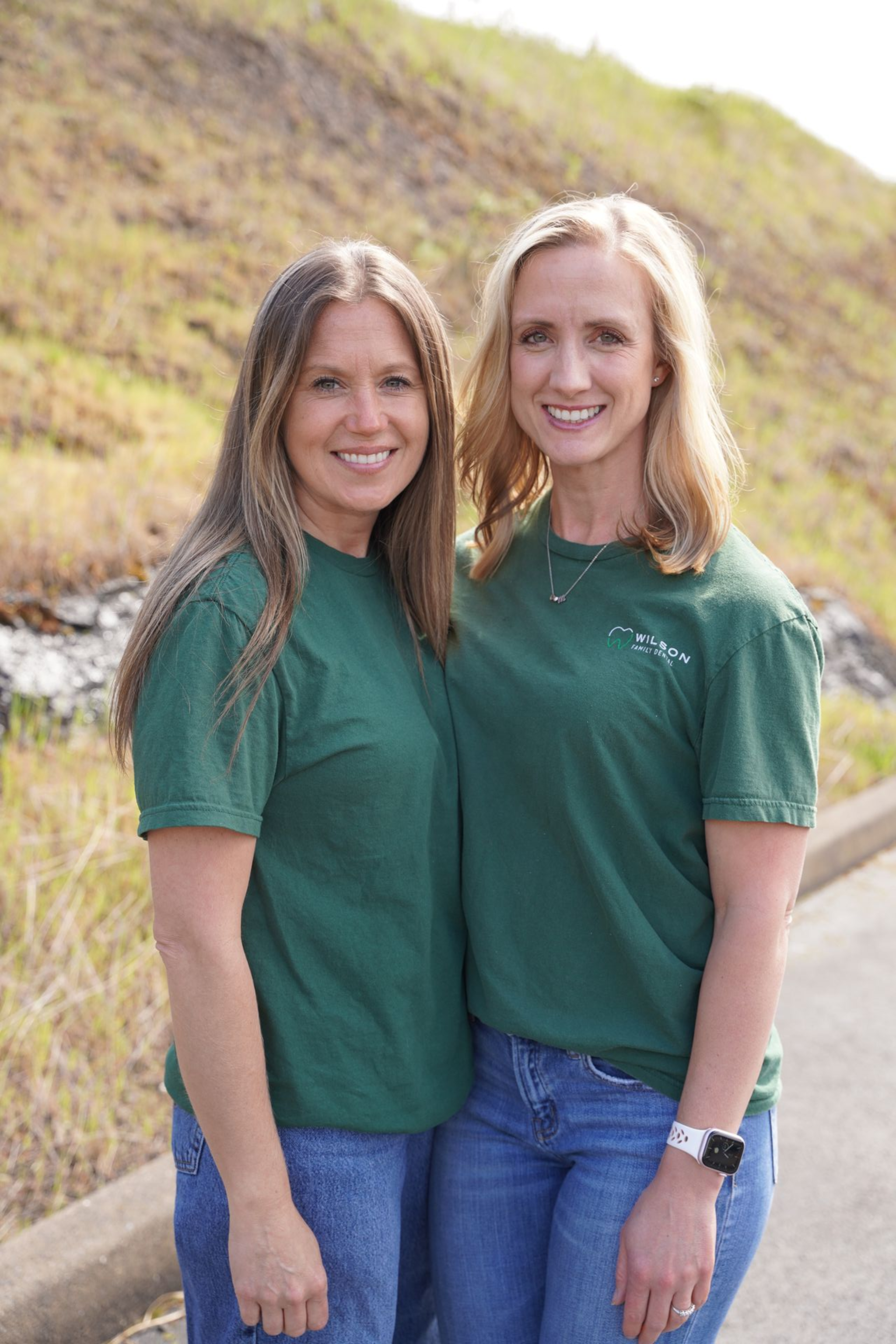 Two smiling people in black jackets stand together outdoors in front of a hillside landscape.
