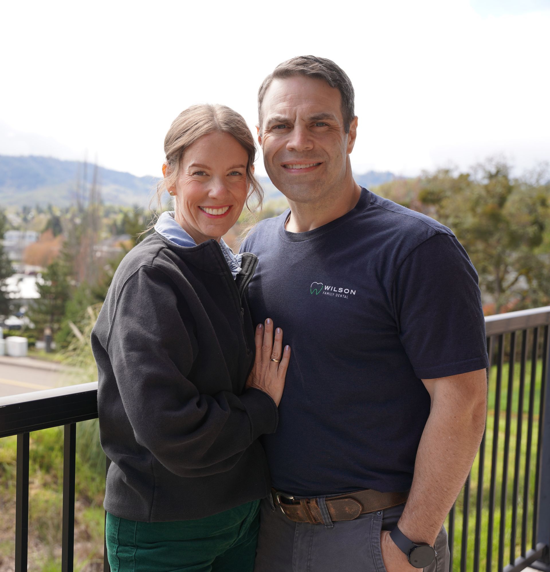 A smiling couple stands outdoors against a background of trees, wearing a dark patterned shirt and a blue dress.