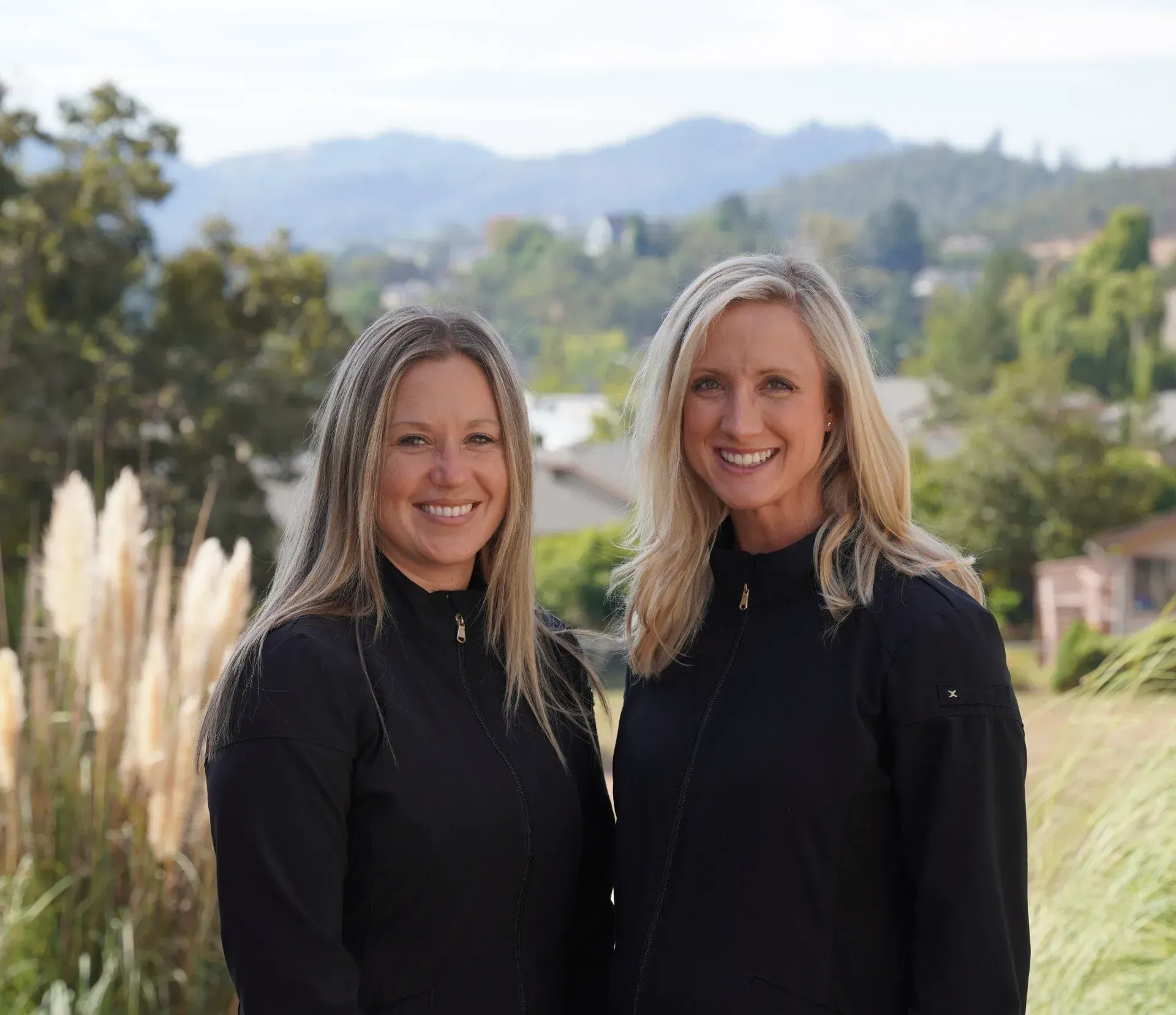 Two smiling individuals wearing black jackets pose side-by-side against a backdrop of green hills and foliage.