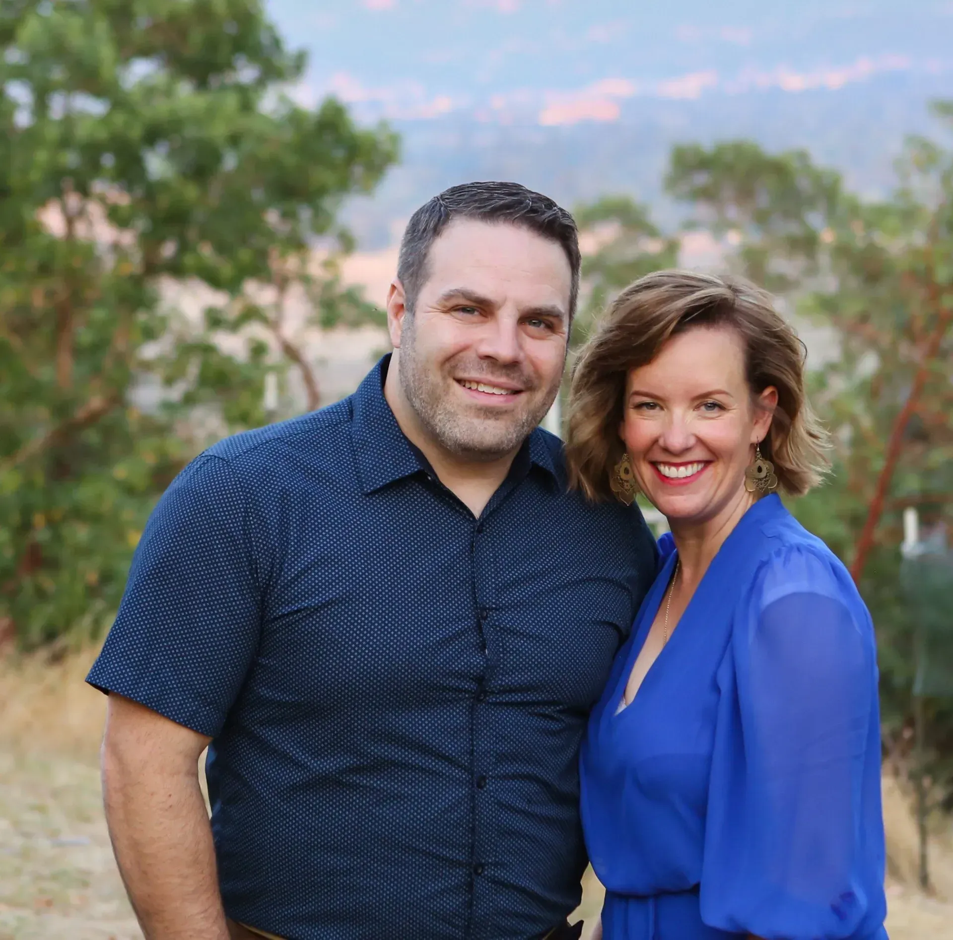 A smiling couple stands outdoors against a background of trees, wearing a dark patterned shirt and a blue dress.