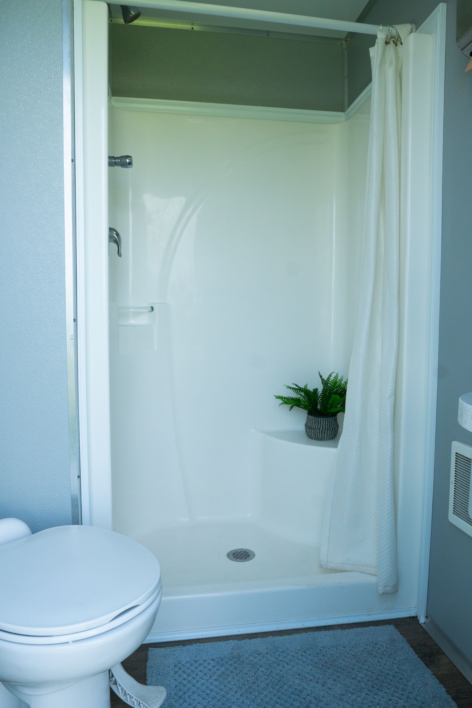 White shower stall and toilet in a small bathroom with a curtain. A small plant sits in the shower.