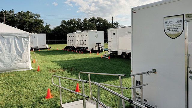 Grassy field with white trailers, orange cones, and a metal ramp near a white tent under a partly cloudy sky