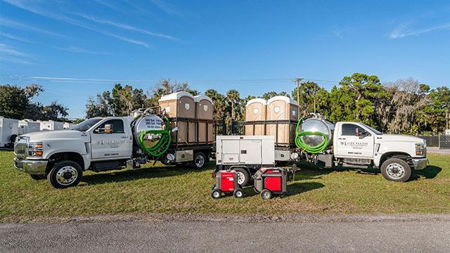 Two white utility trucks with trailers loaded with portable toilets on a grassy field under blue sky