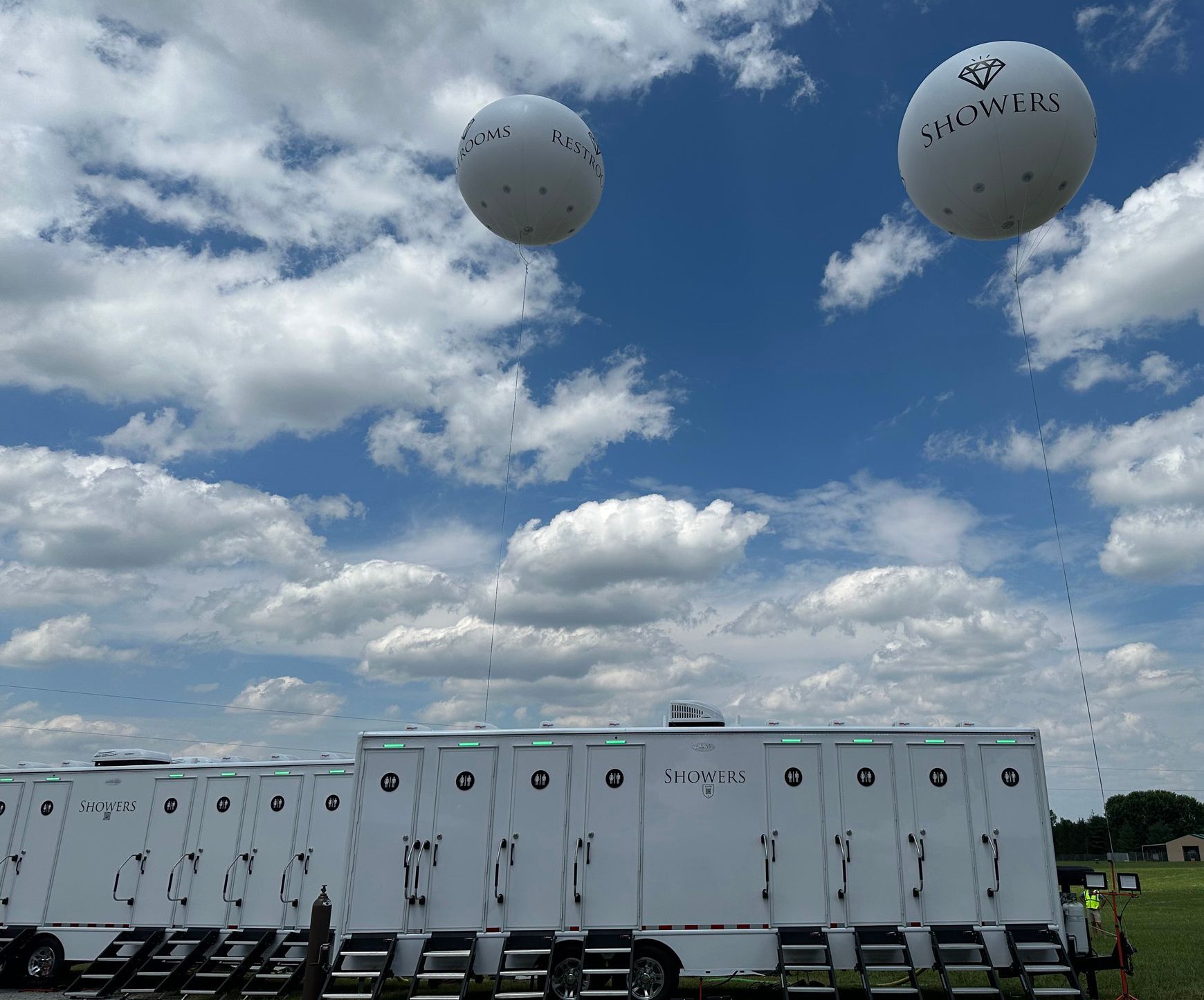 A row of toilet trailers are parked in a field with balloons flying in the sky above them
