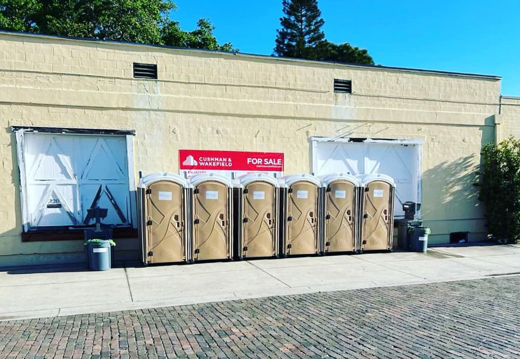 Brown portable toilets lined up in front of a beige building, a