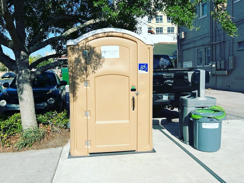 Tan handicap-accessible portable toilet on sidewalk next to a gray trash bin.