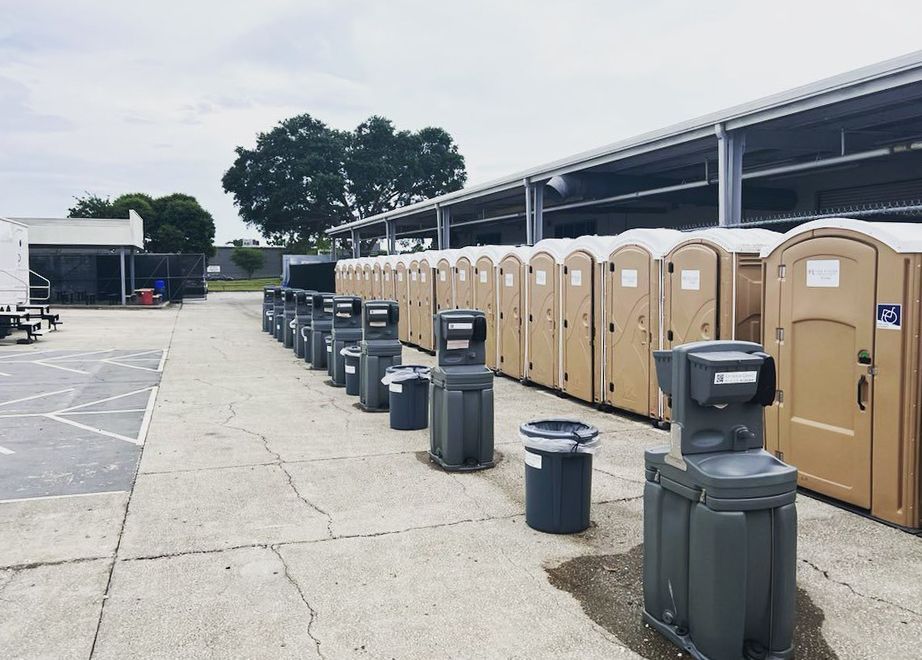 Rows of tan portable toilets and gray handwashing stations on a concrete lot, under a building.