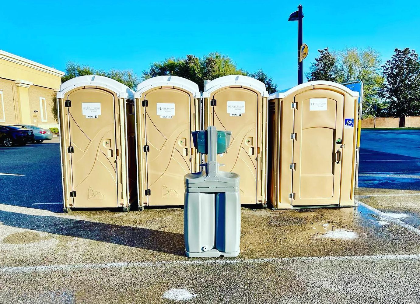 Four tan portable toilets with a hand sanitizer stand in a parking lot. Blue sky and foliage in the background.