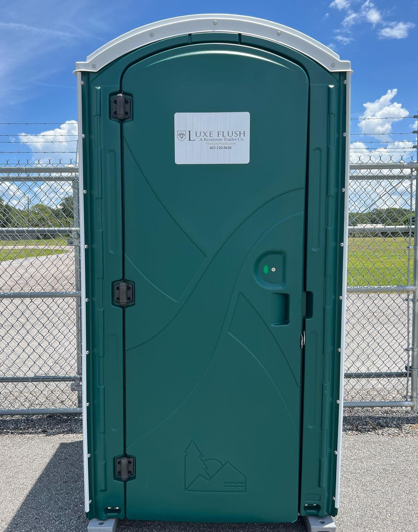 Green portable toilet with closed door, set outside near a chain-link fence and field, blue sky in background.