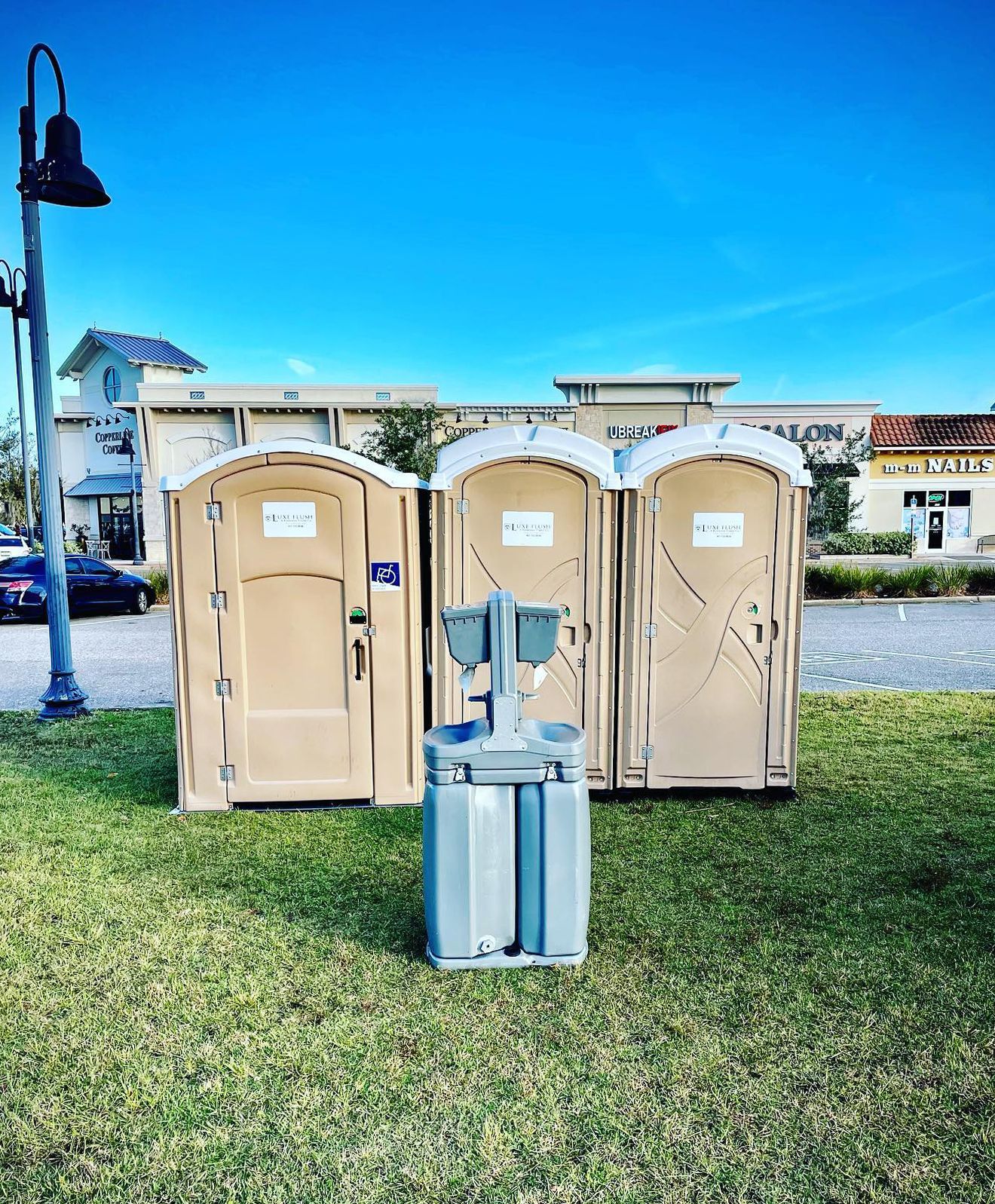 Three beige portable toilets and a handwashing station on grass, with buildings and blue sky in the background.