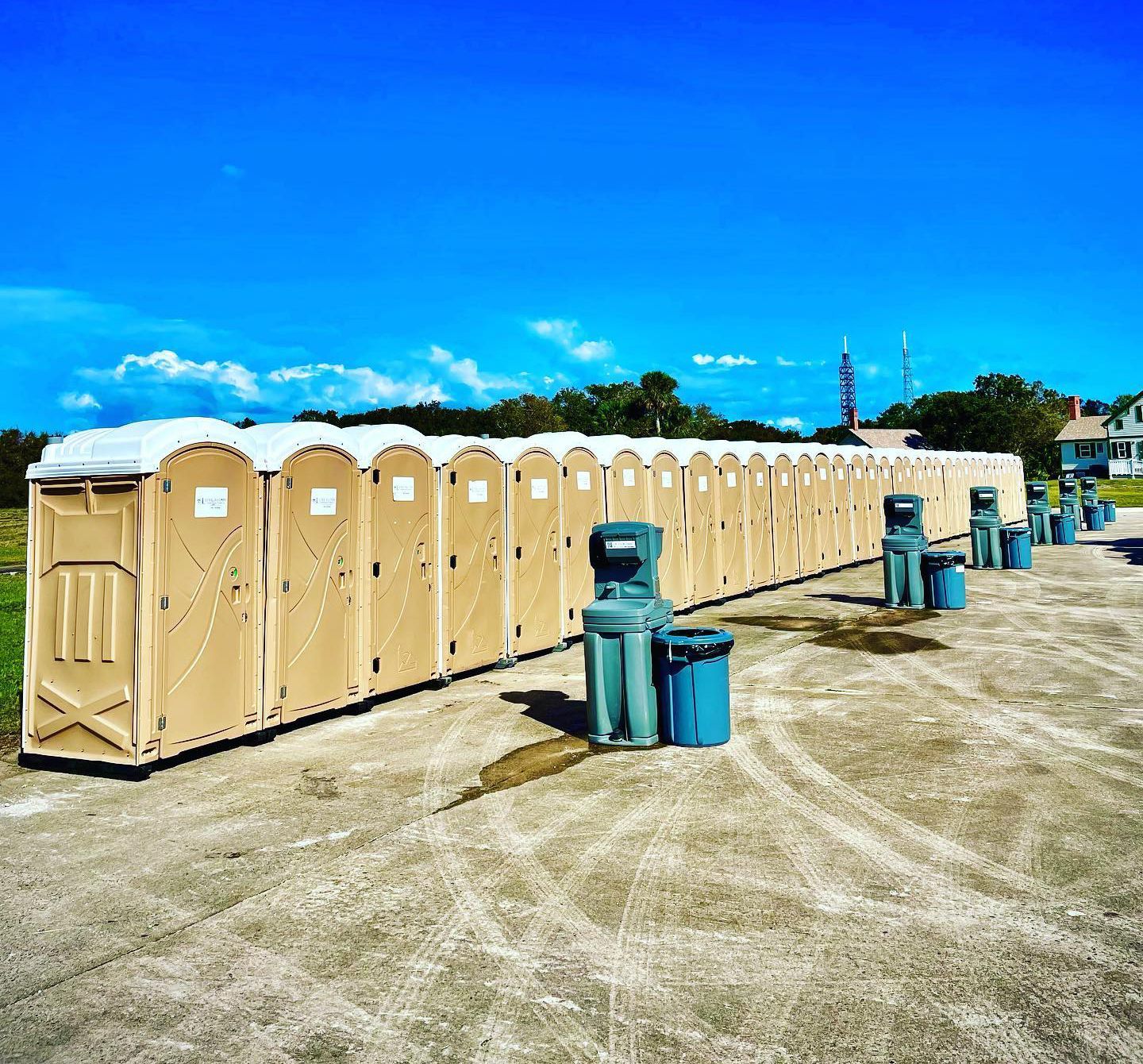 Row of beige portable toilets under a clear blue sky; waste bins in between.