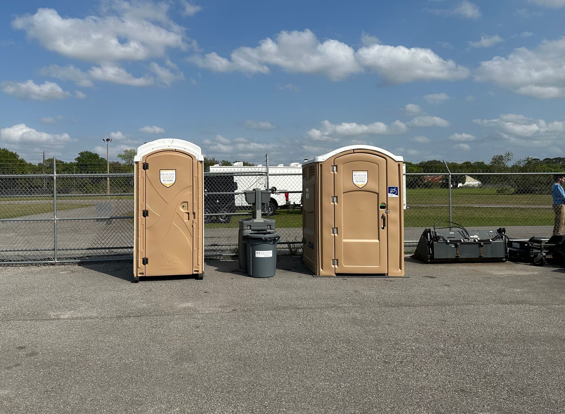 Two tan portable toilets and a hand sanitizer station on asphalt, next to a chain link fence.