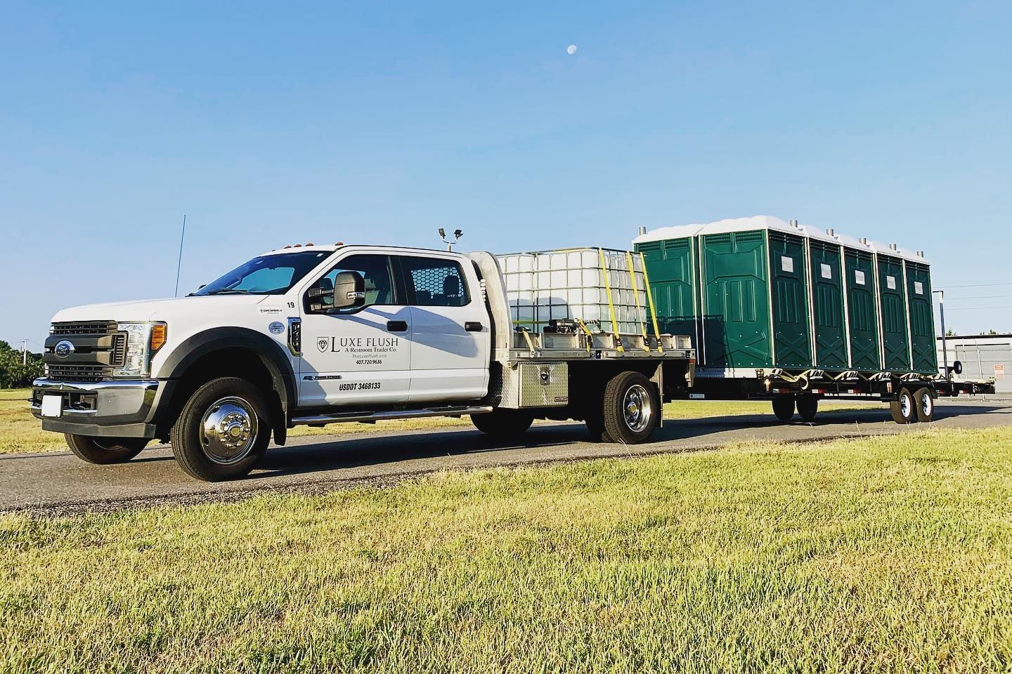 White truck towing a trailer with several green portable toilets on a grassy area under a blue sky.
