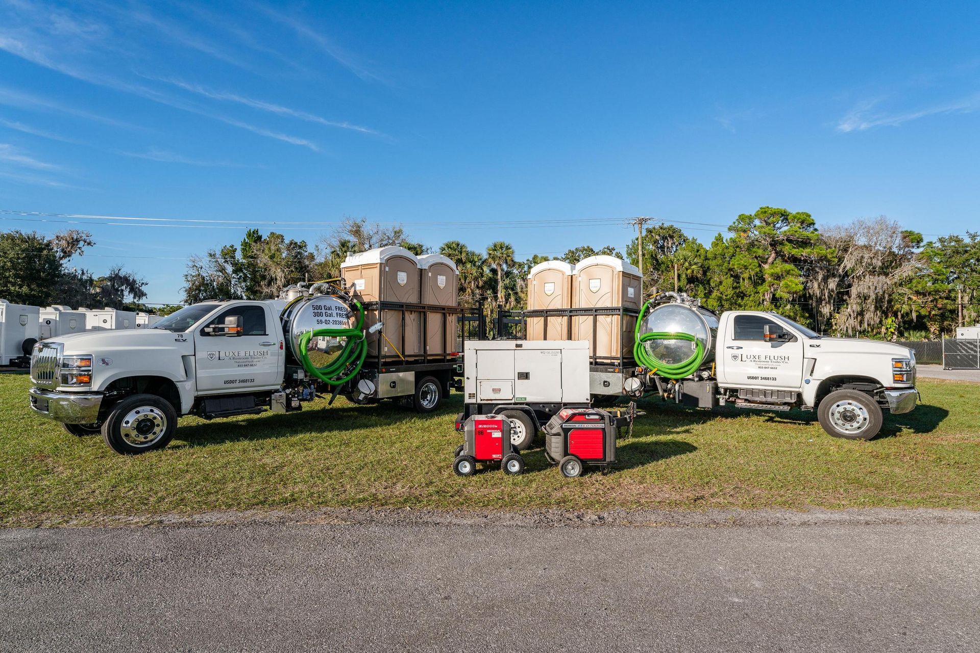 Two white trucks with portable toilets and a generator on a grassy area under a blue sky.