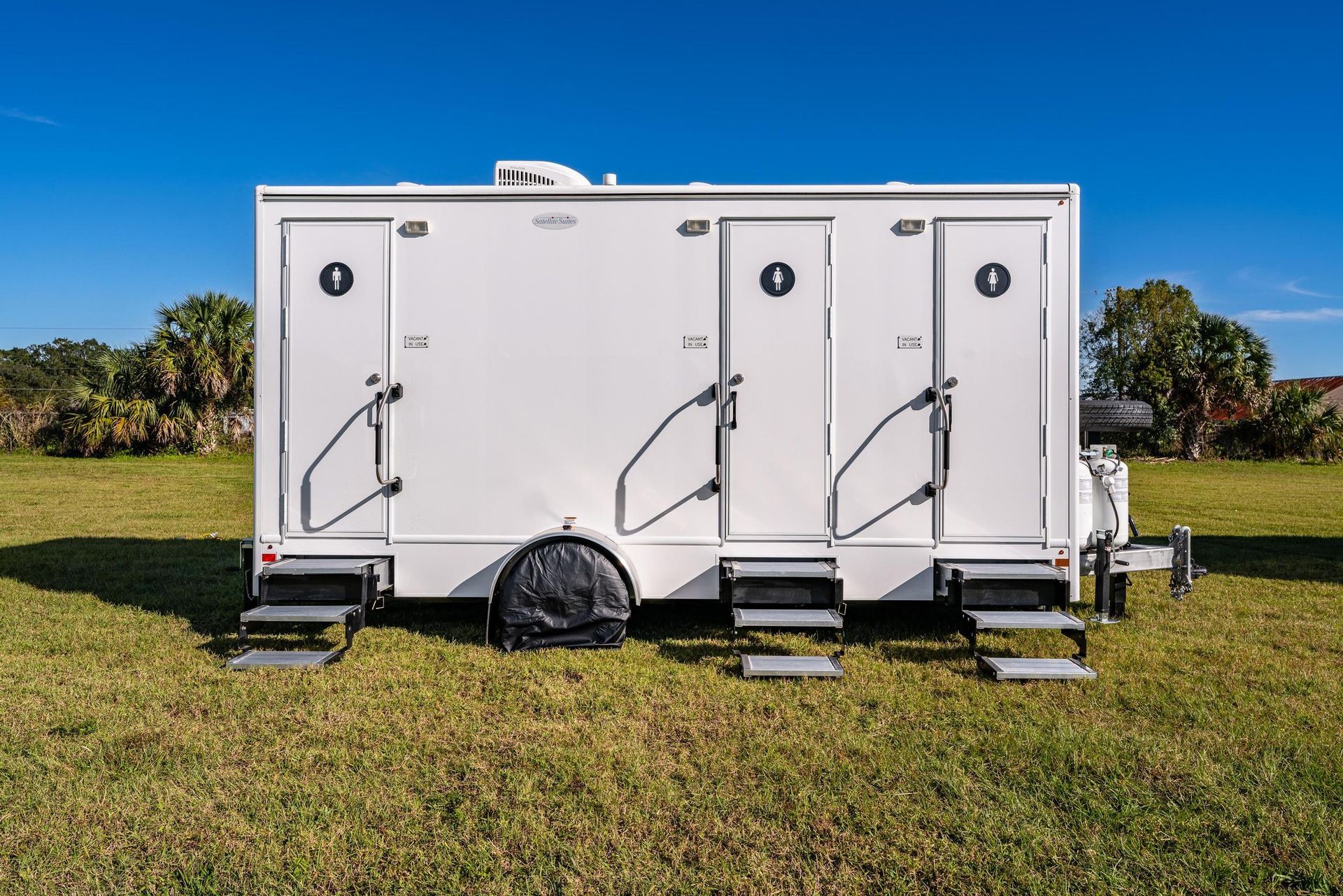 A white trailer with three toilets on it is parked in a grassy field