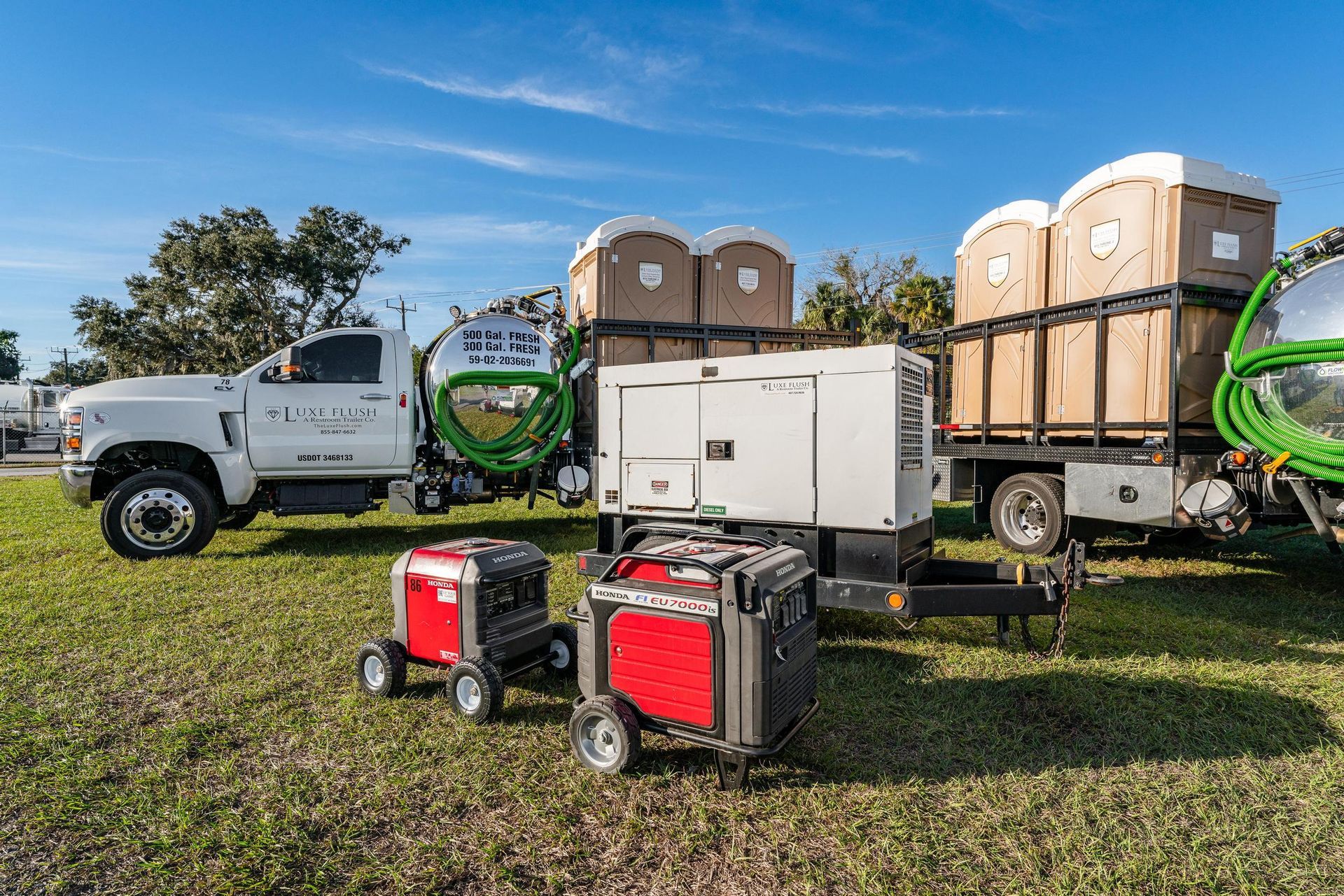 Trucks with portable toilets, generators on grass under a blue sky.