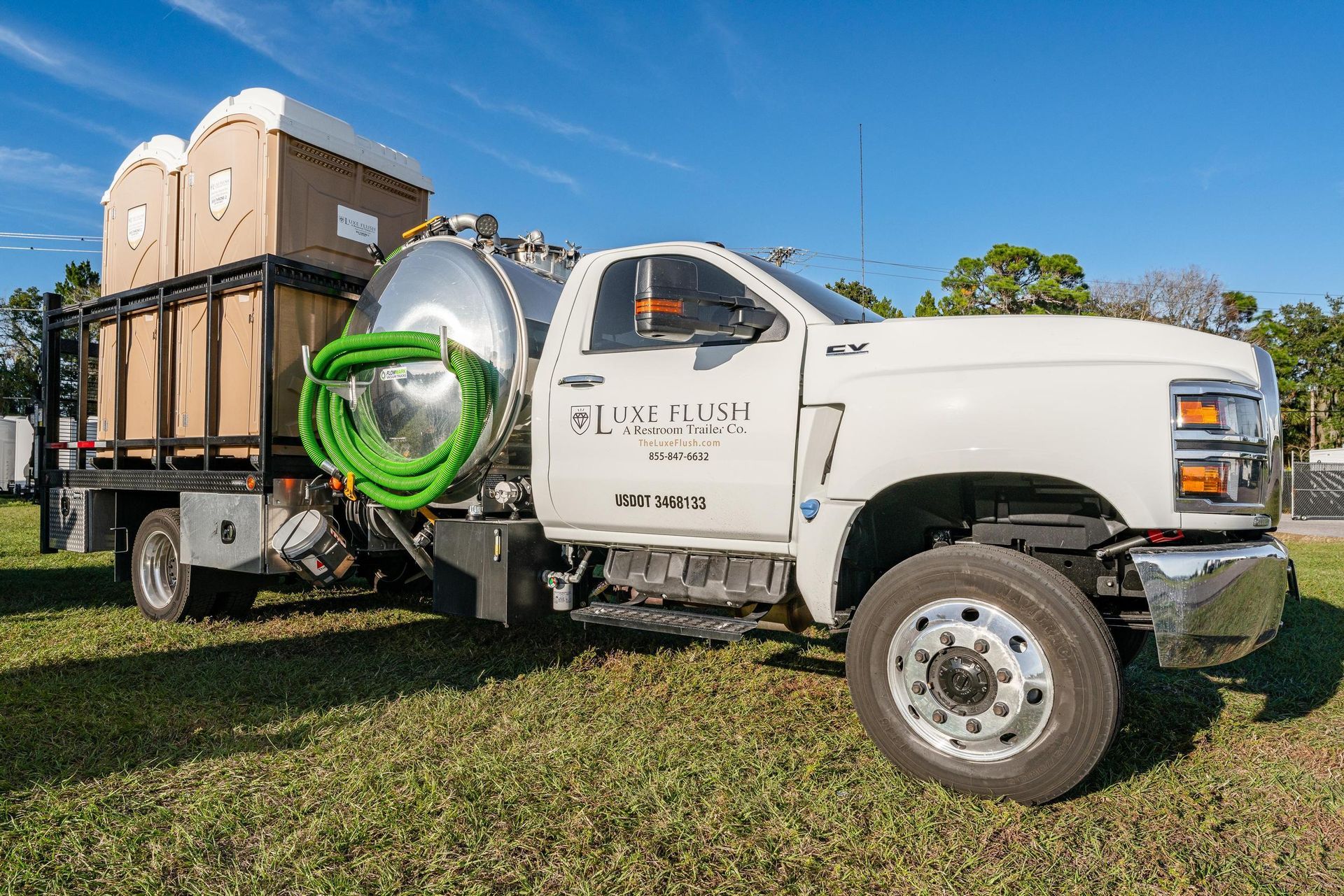 White truck carrying portable toilets and a tank, parked on grass.