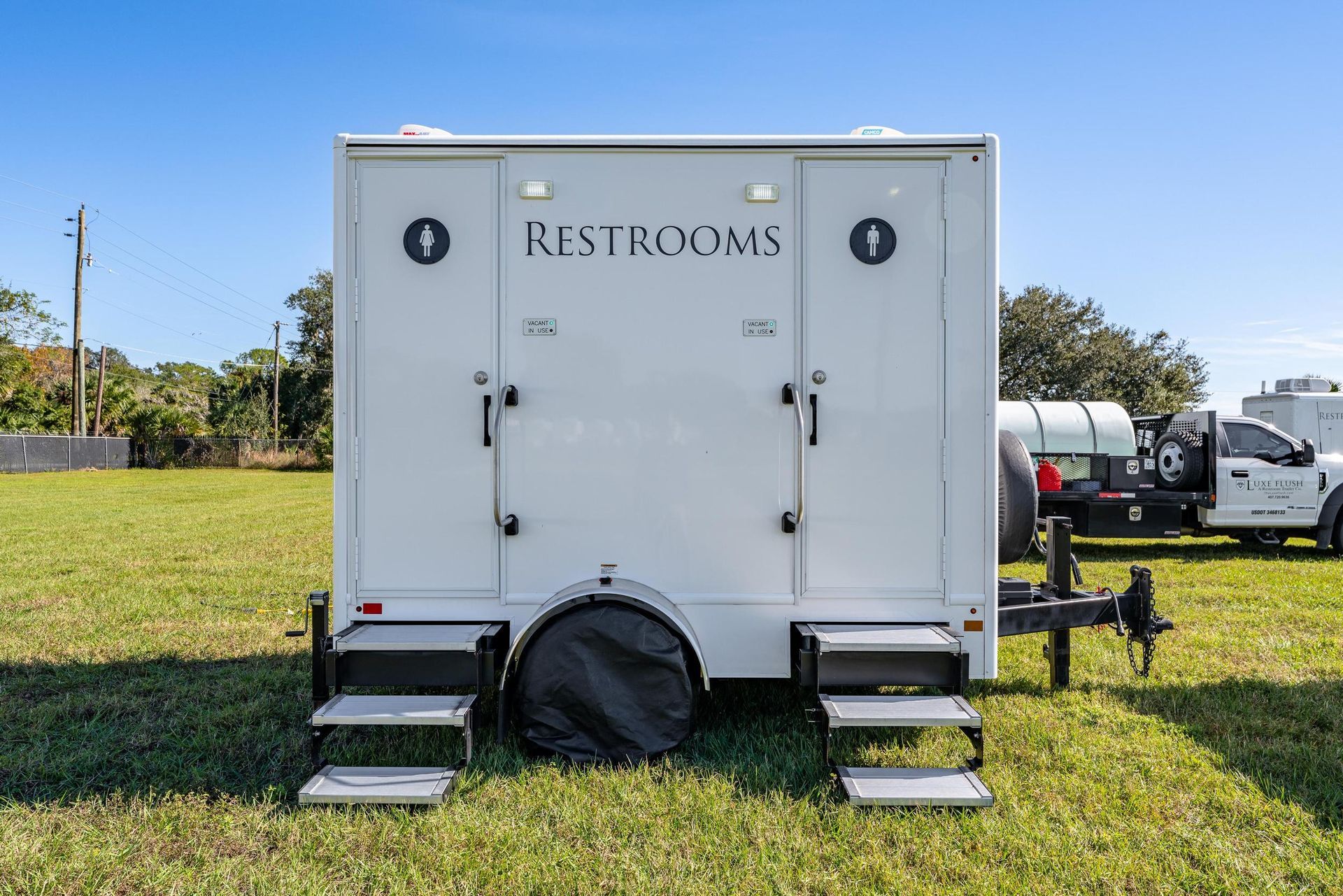 A white restroom trailer is parked in a grassy field