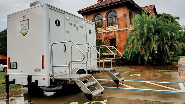 White portable restroom trailer with steps parked outside a brick building in the rain