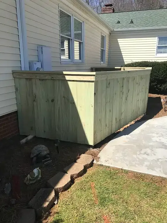 Wooden fence enclosing an outdoor unit next to a house with a concrete patio and green grass.